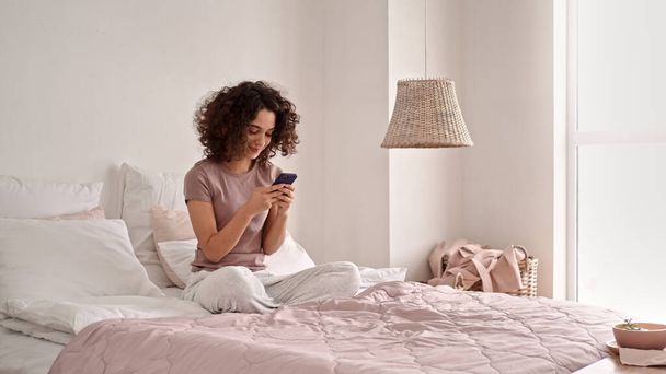 A beautiful shot of Caucasian girl with curly hair smiling and using her phone while sitting on bed - Foto, Bild