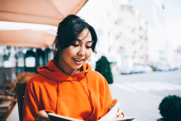 Positive ethnic woman in vivid hoodie with toothy smile looking down while reading book while sitting in cafeteria on street - Foto, imagen