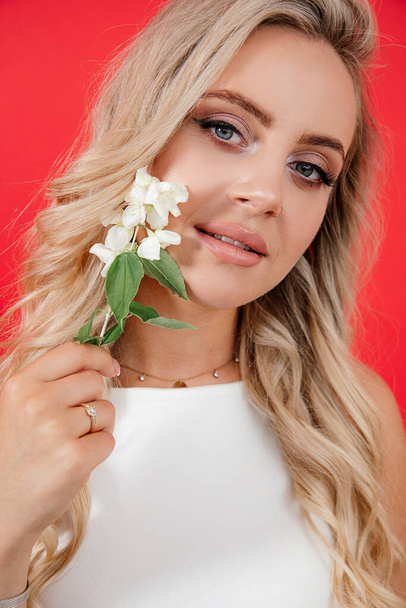 Closeup Portrait of young beautiful blonde woman with jasmine flowers on red background looking at camera. Pure skin, concept of fragrances, natural cosmetics, hair care. Studio shot. - Фото, изображение