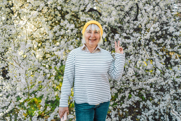 Porträt einer glücklichen schönen blondhaarigen Seniorin in stylischer Kleidung, die im Frühling allein im Park in die Kamera lächelt. Kirschblüte, Allergie - Foto, Bild