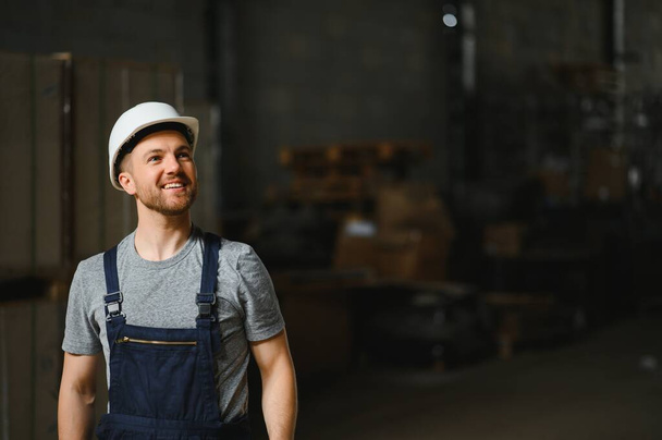 Smiling and happy employee. Industrial worker indoors in factory. Young technician with white hard hat - Фото, изображение