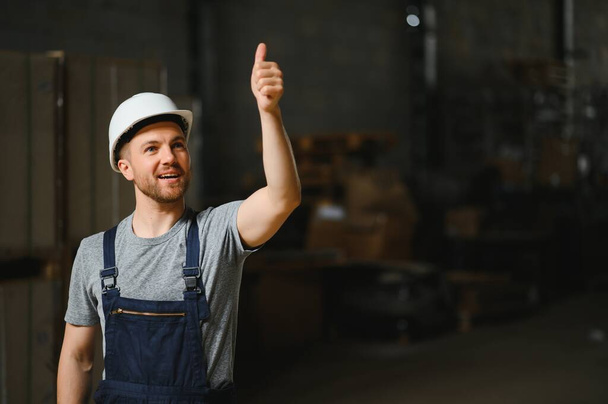Portret van een vrolijke mannelijke arbeider in magazijn met witte helm tussen planken - Foto, afbeelding