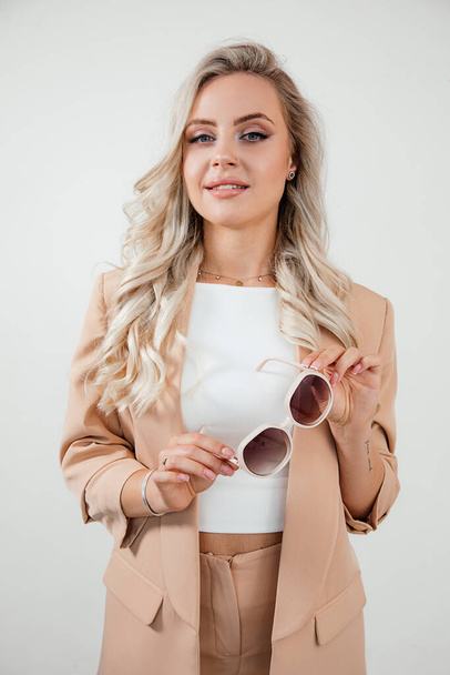 Young beautiful woman, business lady with sunglasses in beige suit on white background studio shot. Optics concept. Blond hair, looking at camera - Foto, Imagem
