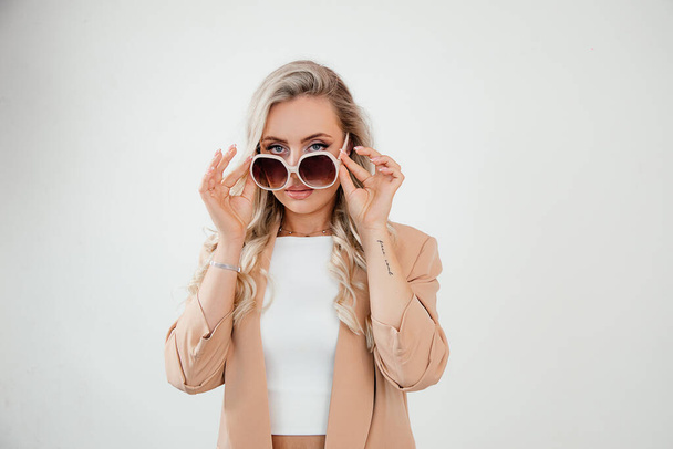 Portrait of Young beautiful woman, business lady with sunglasses in beige suit on white background studio shot. Optics concept. Blond hair, looking at camera - Fotoğraf, Görsel