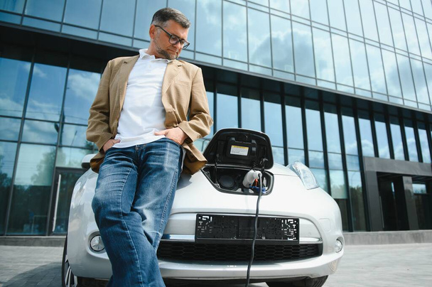 Casual man near electric car waiting for the finish of the battery charging process - 写真・画像