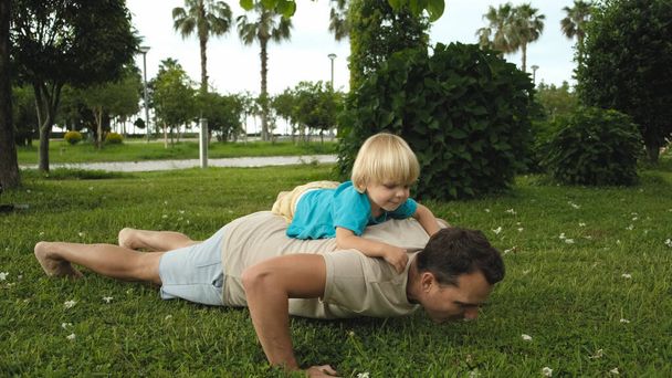 Lapsi kiipeää isien selkään, kun isä punnertaa puistossa. Laadukas 4k kuvamateriaalia - Valokuva, kuva