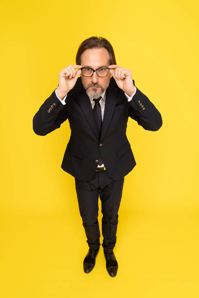 Not happy, serious middle aged business man touching, fixing his eye glasses looking at camera isolated on yellow background. Handsome mature businessman in black suit. Business concept. Copy space. - Фото, изображение