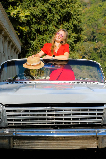 Young happy stylish woman in red dress and hat is standing in vintage car, enjoy summer sunny day and tropical nature. Concept of road travel, vacation, freedom lifestyle, trip - Foto, imagen