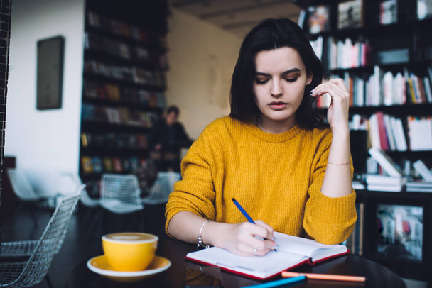 Concentrated young woman in casual clothes writing in planner while sitting at table in modern workplace with cup of hot coffee and creating plan - Foto, Bild