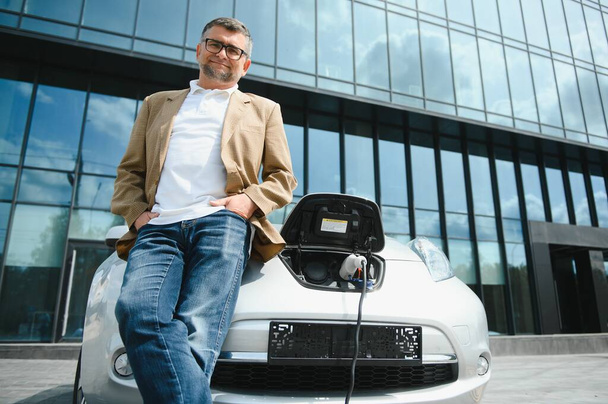 Casual man near electric car waiting for the finish of the battery charging process - Фото, изображение