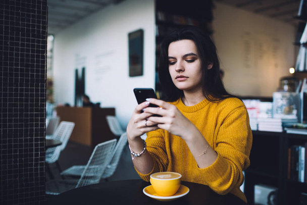 Serious young lady in trendy clothes messaging on mobile phone while sitting at table with coffee and leaning on elbows in modern cafe - Foto, immagini