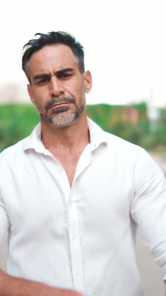 Mature businessman with neat beard wearing white shirt stands with his arms crossed and looks into the camera in the financial district in the city. Successful man looks straight into the camera on modern buildings background - Materiaali, video