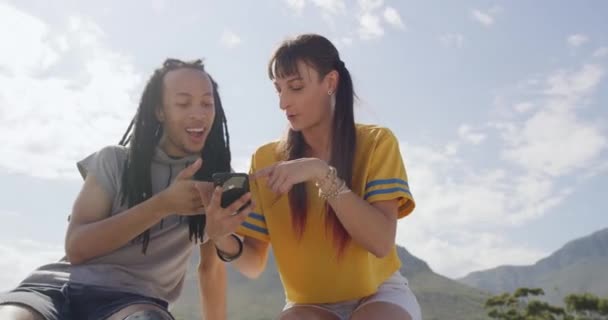 Two young trendy friends using phone and talking at skate park, relaxing with bright copyspace. Modern punks vibing. Diverse couple streaming or checking social media while chilling together outside. - Metraje, vídeo