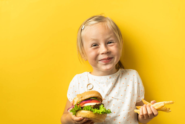 Cute little caucasian girl with blonde hair enjoying burger on a yellow background. Happy kid smiling and eating fast food burger - Foto, Bild