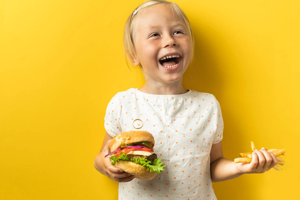 Aranyos kis kaukázusi lány portréja szőke hajjal, aki hamburgert eszik sárga háttérrel. Boldog gyerek gyorskaját eszik. - Fotó, kép