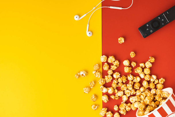 Caramel popcorn scattered from a red and white bucket with remote control on a yellow and red background. Movie or cinema concept. Flat lay. Copy space - Valokuva, kuva