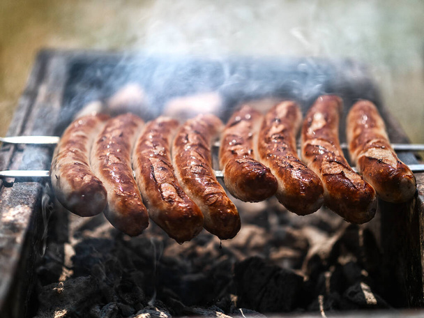Closeup grilled sausages browning on grill outdoors. Meat sausages frying on barbecue grill at backyard. Side view on juicy sausages preparing on round grill outside. - Photo, Image
