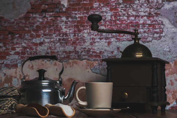 coffee cup with milk jug, grinder and wooden spoons with sugar and ground coffee on a wooden table with a brick wall backdrop - Fotoğraf, Görsel