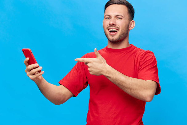 Наслаждался счастливым загорелым красивым мужчиной в красной футболке, указывающим пальцем на телефон, рекомендуемый продукт приложения, позирующего изолированно на синем фоне студии. Копировать макет космического баннера. Концепция образа жизни онлайн-людей - Фото, изображение