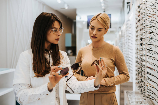Beautiful and fashionable woman choosing eyeglasses frame in modern optical store. Female seller specialist helps her to make right decision. - Foto, immagini