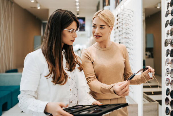 Beautiful and fashionable woman choosing eyeglasses frame in modern optical store. Female seller specialist helps her to make right decision. - Foto, imagen