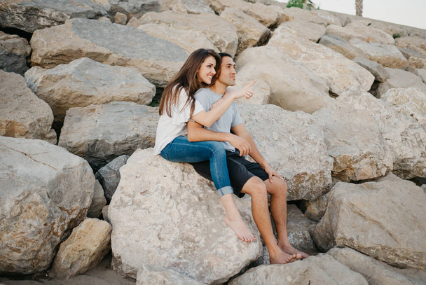 Un hombre hispano está sentado entre las piernas de su novia sonriente en las piedras de un rompeolas en España por la noche. Una mujer feliz está abrazando a su marido en una cita al atardecer en Valencia. - Foto, imagen