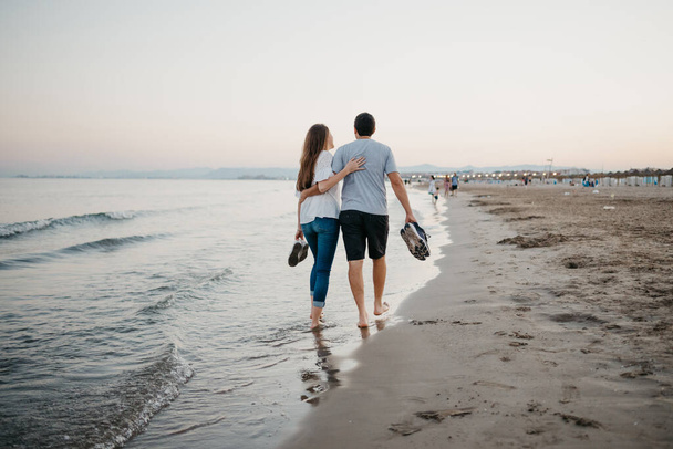 Fotka zezadu muže a ženy, kteří se objímají na písečné pláži bosí za soumraku ve Španělsku. Pár turistů na rande u Baleárského moře ve Valencii. - Fotografie, Obrázek