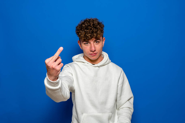 Young spanish man wearing white sweatshirt with curly hair standing over isolated blue background Showing middle finger, impolite and rude fuck expression - Fotoğraf, Görsel