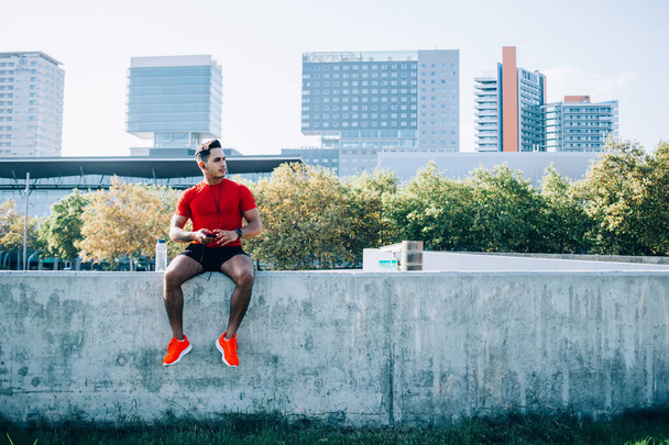 Serious ethnic young man in sportswear with earphones preparing for jogging using smartphone for listening music sitting on stone wall in stadium - Photo, Image