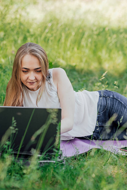 Op het spandoek werkt een jong meisje met een laptop in de frisse lucht in het park, zittend op het gazon. Het concept van werken op afstand. Werk als freelancer. Het meisje neemt cursussen op een laptop en glimlacht - Foto, afbeelding