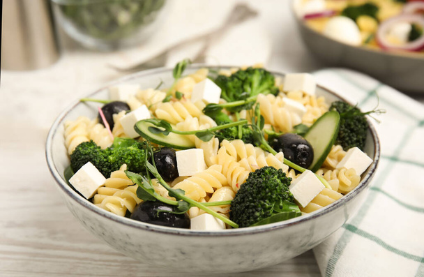 Bowl of delicious pasta with cucumber, olives, broccoli and cheese on white wooden table, closeup - 写真・画像