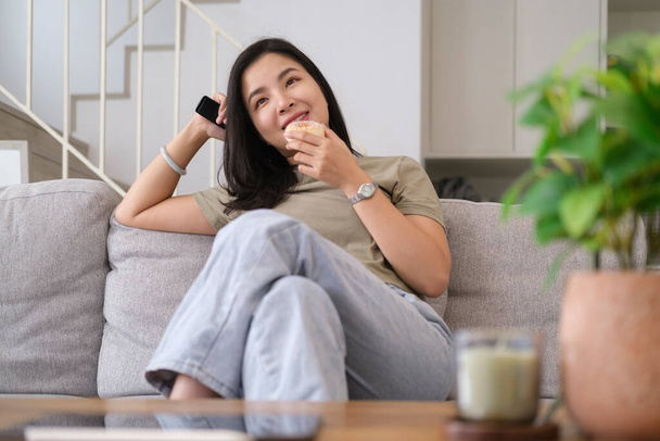 Smiling young woman relaxing on comfortable sofa, enjoying watching television at home. - 写真・画像