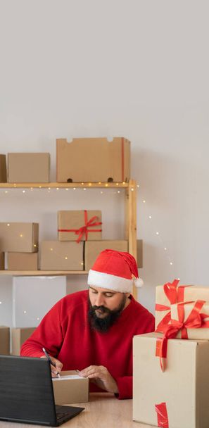 Noel Baba şapkası takan Hintli sakallı adam bir kutu dolusu kutuda çalışır ve Noel için internetten hediye alır. Küçük işletme sahibi mutlu yıllar satıyor. dikey pankart - Fotoğraf, Görsel