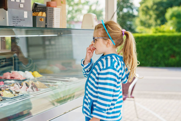 Schattig klein peuter meisje kiezen en kopen van ijs in outdoor ijs truck cafe. Gelukkige kleuter met een bril die naar verschillende soorten ijs kijkt. Zoet zomerdessert. - Foto, afbeelding