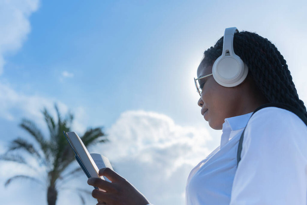 Африканська жінка у навушниках, читаючи книжку на відкритому повітрі, не звертає на це уваги. Вибіркове фокусування. Життєпис і концепція дозвілля. - Фото, зображення