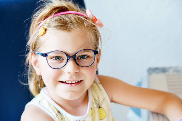 Portrait of a cute preschool girl with eye glasses indoors. Happy funny child wearing new blue glasses - Fotó, kép