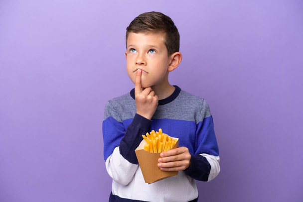 Kleiner Junge mit frittierten Chips isoliert auf lila Hintergrund mit Zweifeln beim Blick nach oben - Foto, Bild