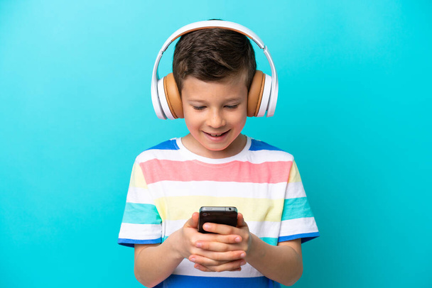 Little boy isolated on blue background listening music and looking to mobile - Photo, Image