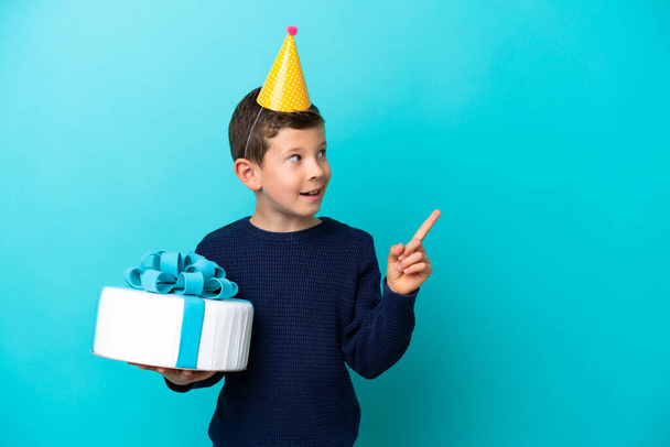 指を持ち上げながら解決策を実現しようとしている青の背景に隔離された誕生日ケーキを保持する小さな男の子 - 写真・画像