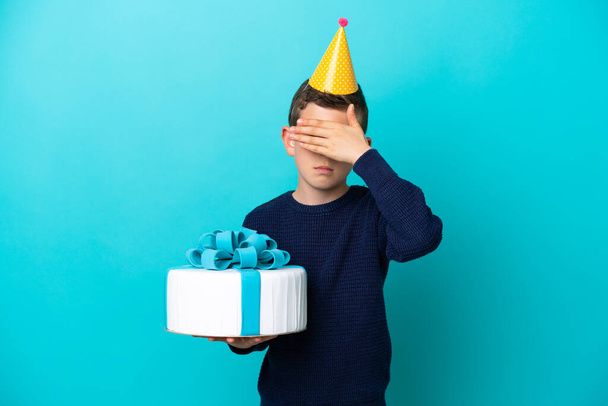 Little boy holding birthday cake isolated on blue background covering eyes by hands. Do not want to see something - Zdjęcie, obraz