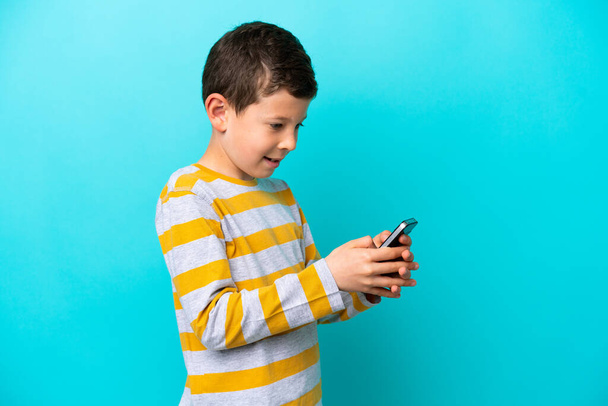 Kleine jongen geïsoleerd op blauwe achtergrond verzenden van een bericht of e-mail met de mobiele - Foto, afbeelding