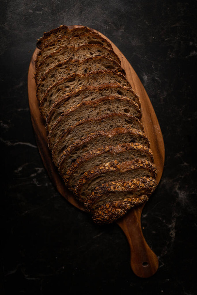 A top view of a sliced rye bread on a wooden board on a black table - Фото, изображение