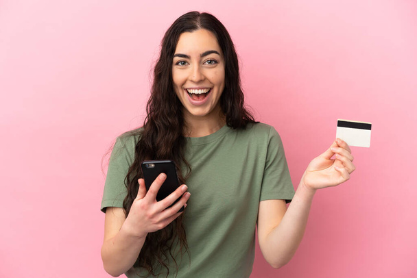 Молодая белая женщина, изолированная на розовом фоне, покупает мобильный телефон и держит кредитную карту с удивленным выражением лица - Фото, изображение