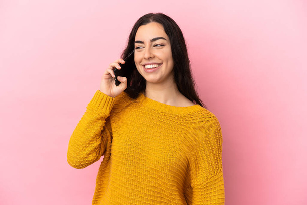 Pembe arka planda izole edilmiş cep telefonu kullanan genç beyaz kadın yana bakıyor ve gülümsüyor. - Fotoğraf, Görsel