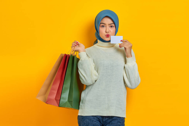 Cheerful beautiful Asian woman in white sweater holding shopping bags and credit card isolated over yellow background - Foto, Imagen