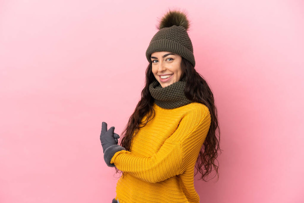 Νεαρό καυκάσιο κορίτσι με χειμερινό καπέλο απομονωμένο σε μωβ φόντο που δείχνει πίσω - Φωτογραφία, εικόνα