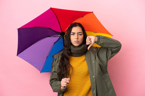 Mladá žena drží deštník izolovaný na růžovém pozadí ukazující palec dolů s negativním výrazem - Fotografie, Obrázek