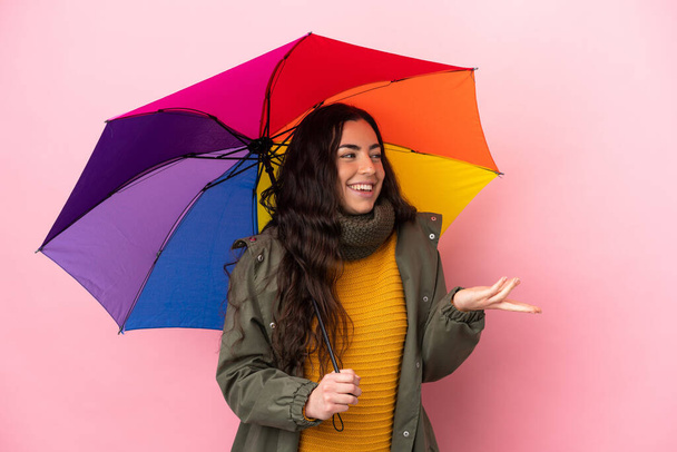 Young woman holding an umbrella isolated on pink background with surprise expression while looking side - Fotografie, Obrázek