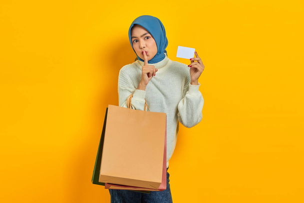 Krásná asijská žena v bílém svetru drží nákupní tašku a kreditní kartu, aby ticho gesto izolované přes žluté pozadí - Fotografie, Obrázek
