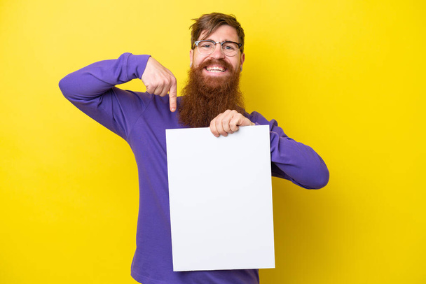 Zrzavý muž s vousy izolovaný na žlutém pozadí drží prázdný štítek se šťastným výrazem a míří na něj - Fotografie, Obrázek